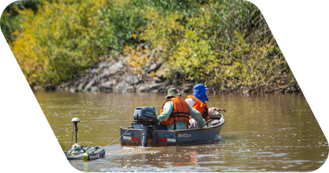 Dois homens em um barco num rio. Ambos estão de costas para foto e usam colete e chapéu. Ao fundo, há uma vasta vegetação.