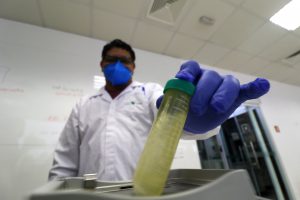 Homem segurando um tubo de ensaio com um líquido dentro. Ele usa jaleco, máscara, luvas e óculos e está em laboratório.