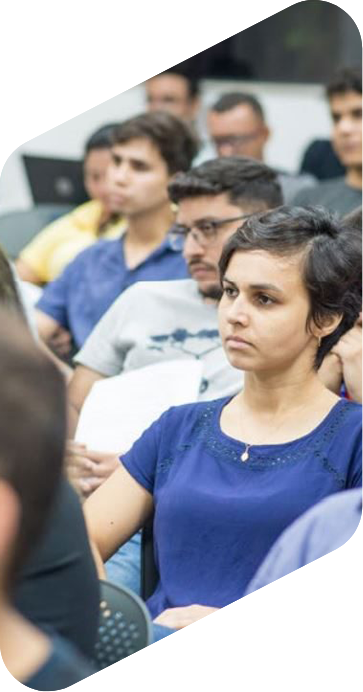 Mulher, de cabelos curtos, em uma sala de aula olhando fixamente para frente. Ela usa blusa azul escura e colar. Atrás dela, há vários alunos sentados.