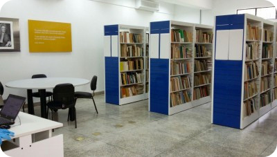 Prateleiras brancas e azul escuro da biblioteca ITV Desenvolvimento Sustentável com diversos livros. Na lateral esquerda, há uma mesa branca com 3 cadeiras e dois quadros na fixados na parede.