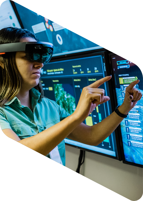 Mulher com óculos de realidade virtual gesticulando com as mãos. Na frente dela, há quatro televisores ligados com conteúdos diferentes