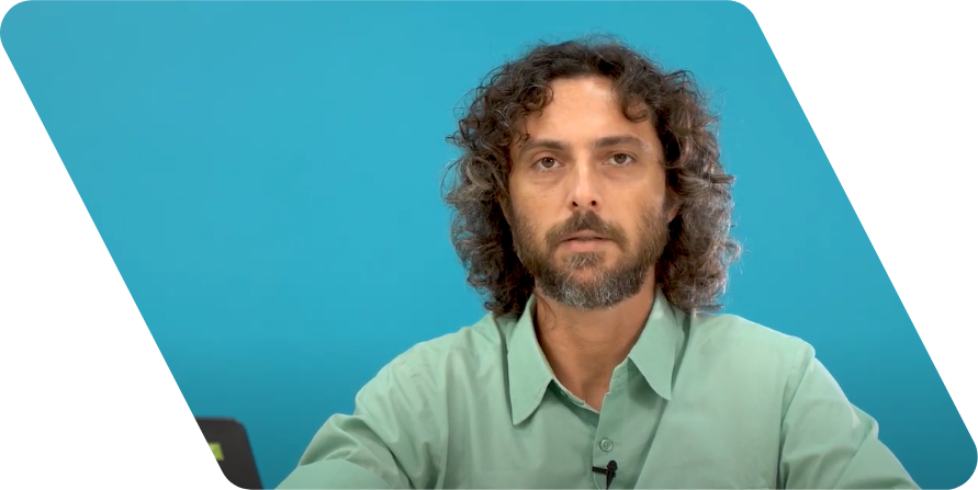 Homem branco de cabelos castanhos enrolados até a altura dos ombros e barba olhando para a câmera. Ele usa uma camisa de cor verde clara e está na frente de uma parede azul.