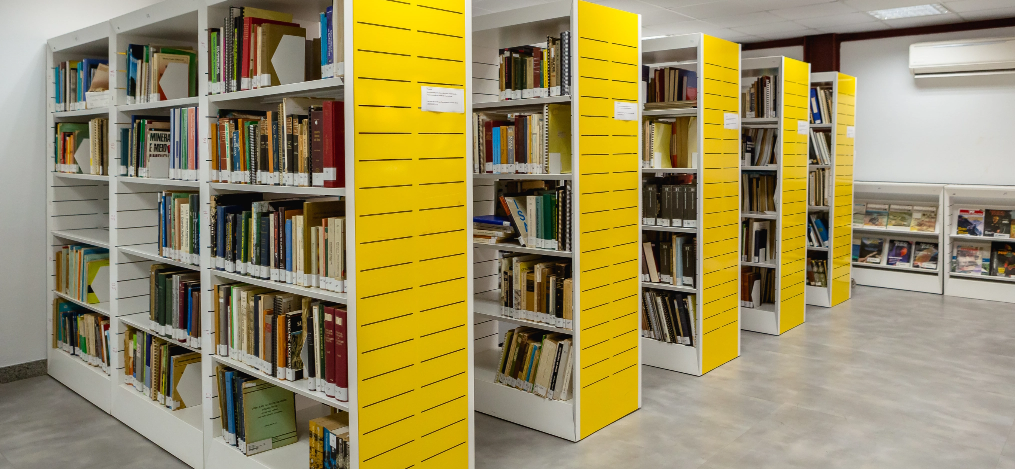 Corredor da biblioteca ITV Mineração com cinco prateleiras nas cores amarela e branca repletadas de livros.
