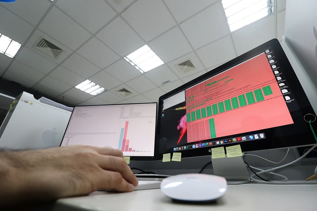 Mão de uma pessoa usando um mouse em frente a dois monitores de computador que exibem gráficos de dados e código.