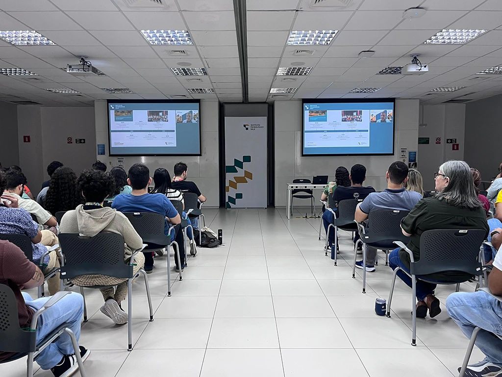 Grupo de pessoas assistindo a uma apresentação com duas telas de projeção em uma sala de conferência.